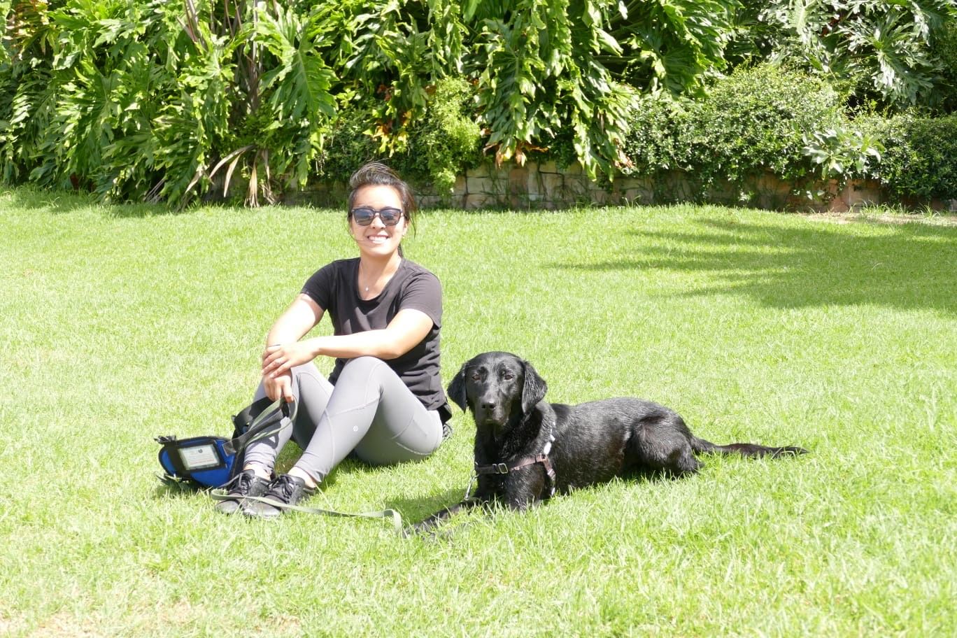 Diona sitting on the grass in the sun with Murphy, her black Labrador assistance dog, settled beside her